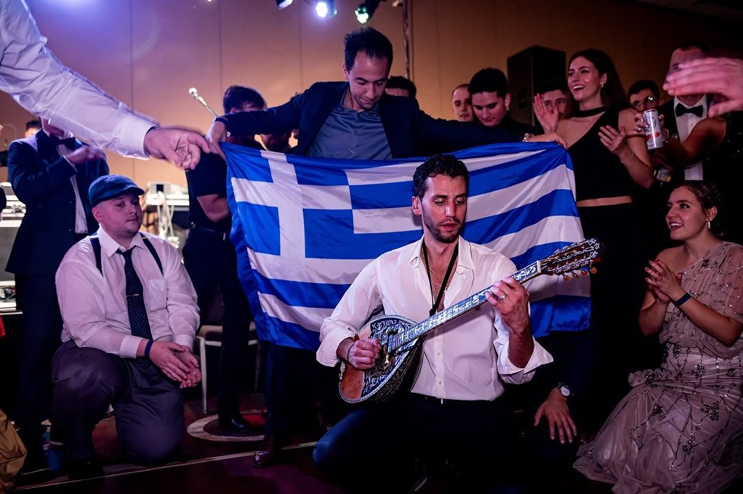 Oneiro musician playing bouzouki beside a Greek flag at a party